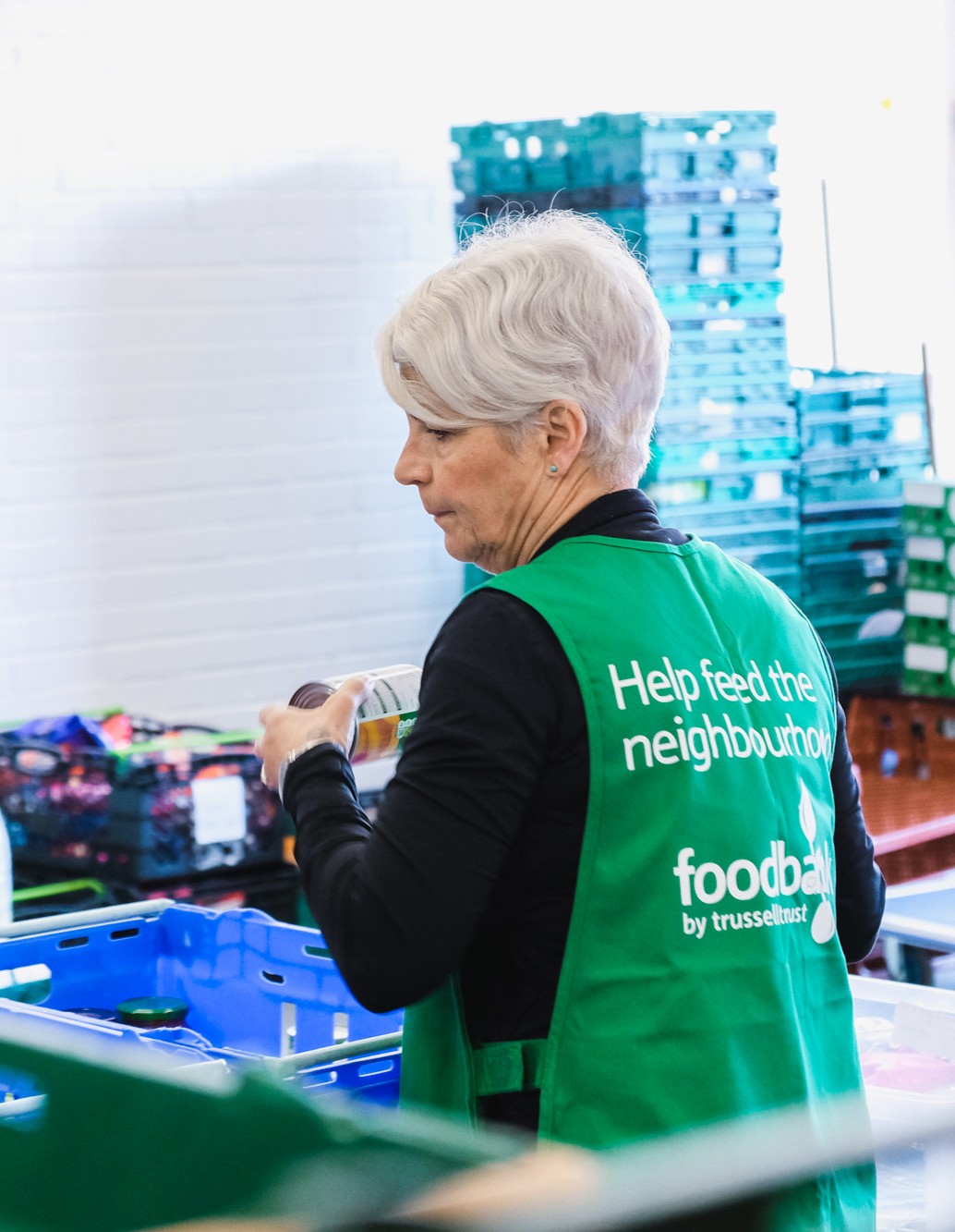A volunteer packs a crate, she is turned away from the camera, her tabard reads Help Feed the Neighbourhood