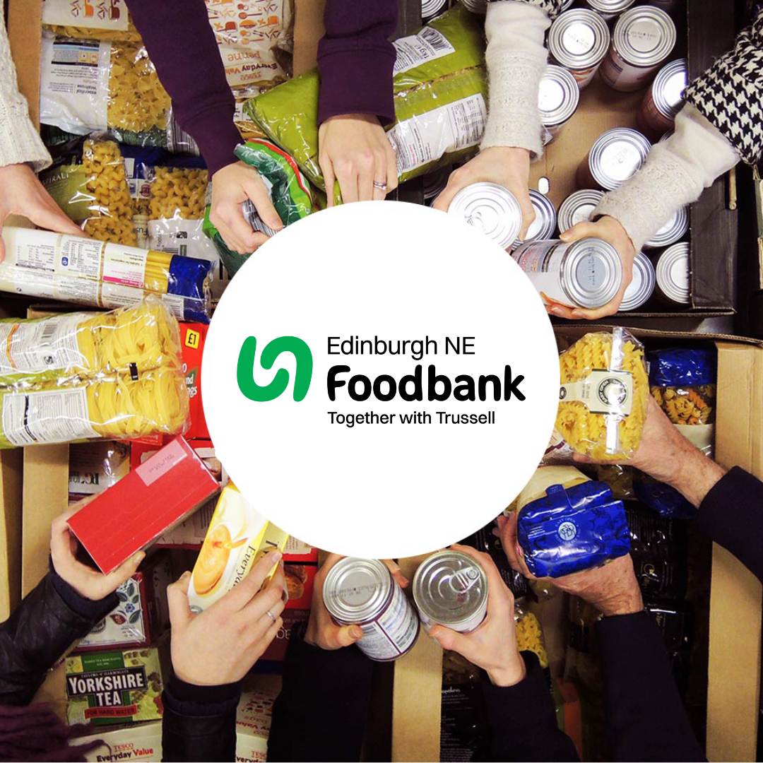 Top down image of lots of hands holding items of food like tins and pasta in a circle, in front of boxes and crates of more food.